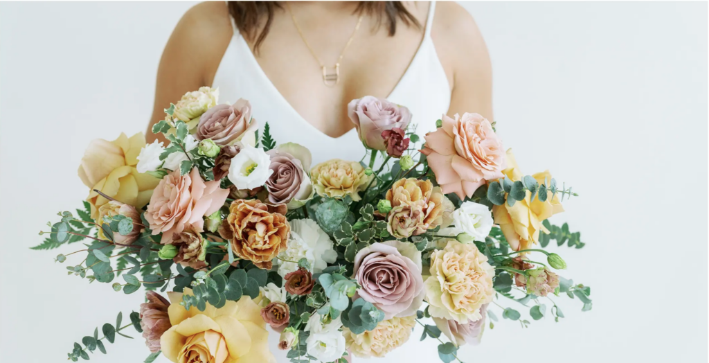 White woman holding an arrangement of white, yellow, pink, and purple roses with a variety of greenery and assorted flowers.