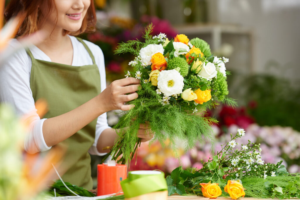 Person arranging a bouquet of yellow, white, and green flowers and ferns at a flower shop.