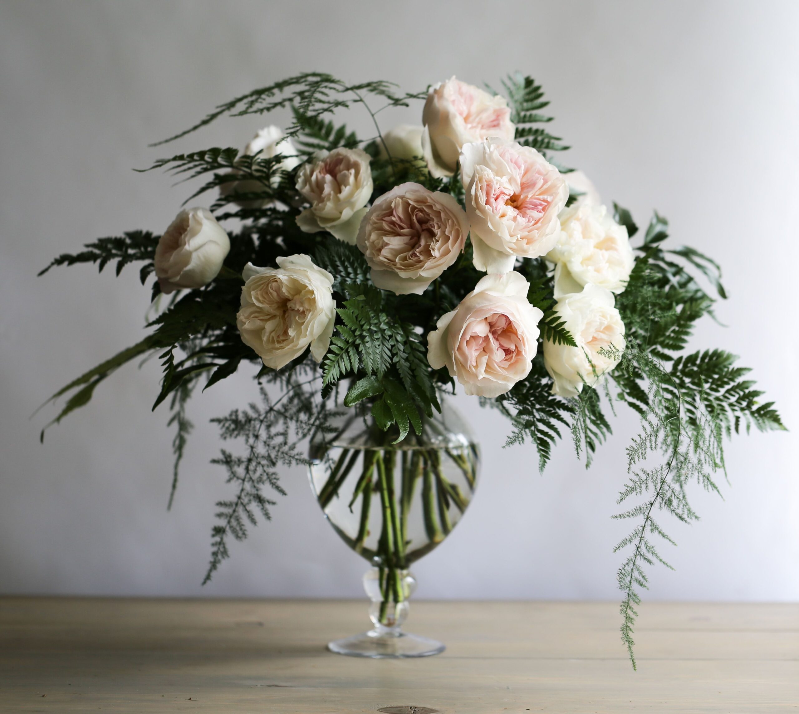 Floral arrangement of cream-colored roses with pink centers and Plumosus fern leaves in a glass vase, placed on a wooden surface with a plain white background.