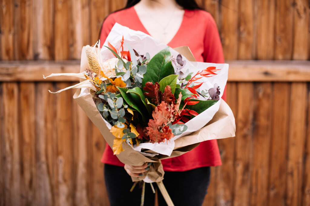 A woman in a red shirt is holding a bouquet of autumn-themed flowers and greenery, with a wooden fence in the background.