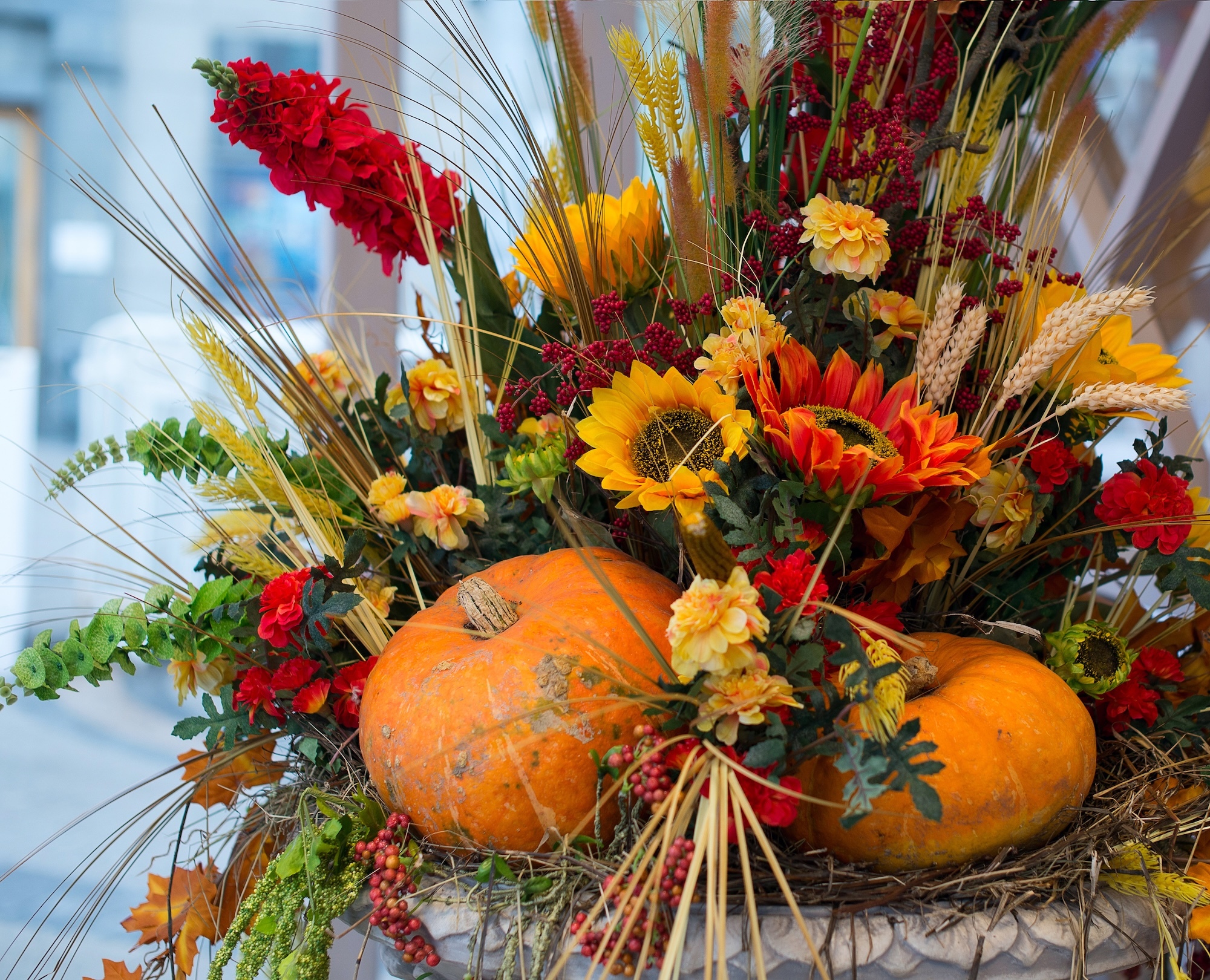  A vibrant autumn floral arrangement featuring bright orange pumpkins, sunflowers, red and yellow flowers, wheat stalks, and greenery, creating a festive harvest display.