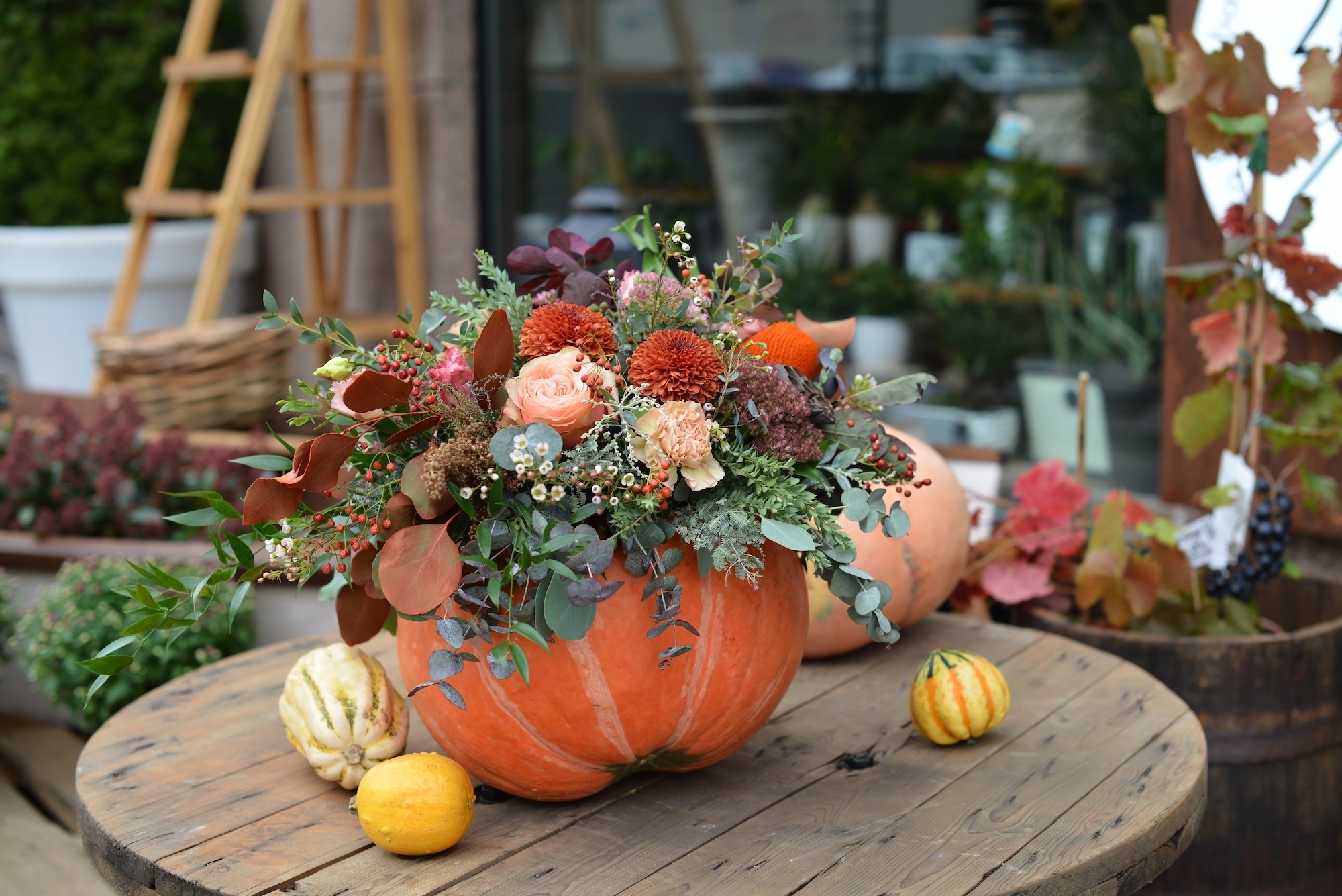 A creative autumn centerpiece featuring a large pumpkin used as a vase, filled with orange roses, chrysanthemums, greenery, and small decorative gourds on a rustic wooden table.