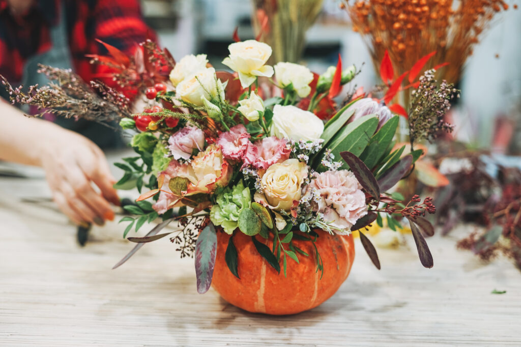 A vibrant floral arrangement in a carved pumpkin vase featuring roses, carnations and various autumnal greenery. A person in the background is arranging the flowers on a light wooden table.