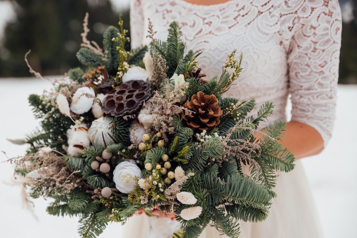 A close-up of a winter bouquet held by a person in a lace dress, featuring evergreen branches, pinecones, cotton stems, dried flowers and lotus seed pods.