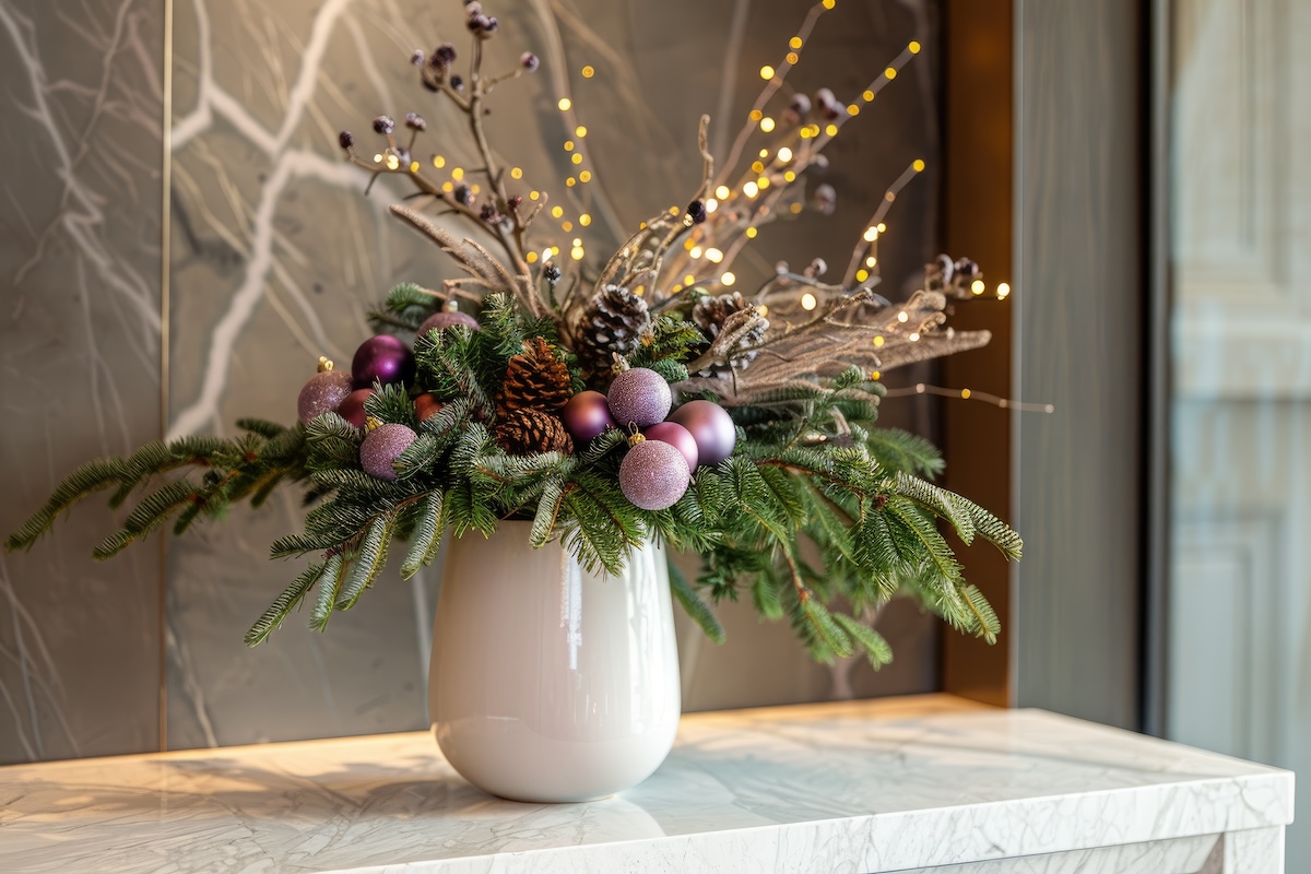 A festive holiday arrangement in a white vase on a marble surface, adorned with evergreen branches, pinecones, purple ornaments and twinkling fairy lights.